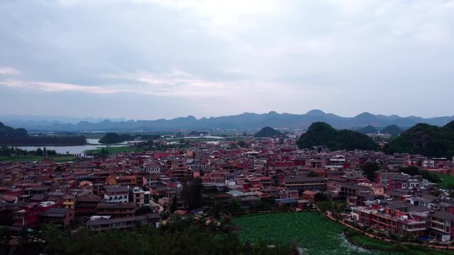 An aerial panoramic view of a dense traditional village nestled among karst mountains with water bodies nearby under an overcast sky.