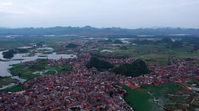 Aerial panorama of a lakeside town surrounded by karst hills, fields and water bodies under an overcast sky.