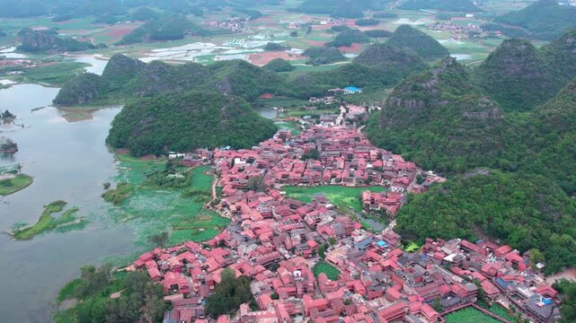 An aerial shot shows a village with red rooftops nestled among karst mountains and water bodies in a scenic rural landscape.