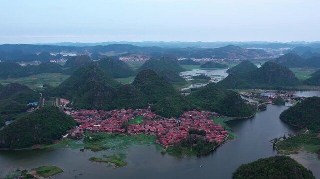 An aerial view of a red-roofed village surrounded by green karst hills and winding waterways in a natural landscape.