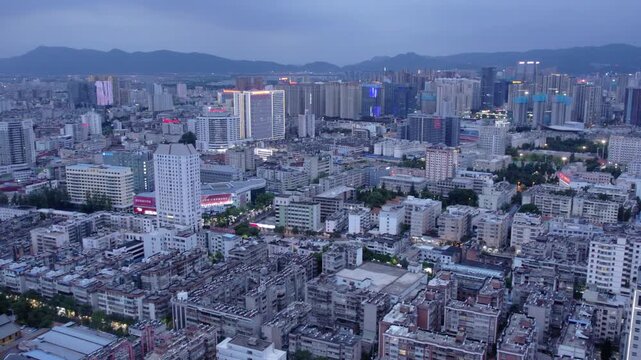 An aerial panoramic view of a dense urban city with mixed buildings, backed by distant mountains during dusk.