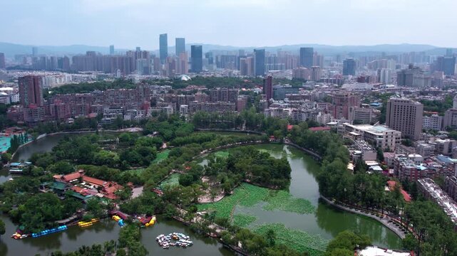 A high-angle aerial shot shows an urban park with a lake, surrounded by city buildings and green trees.