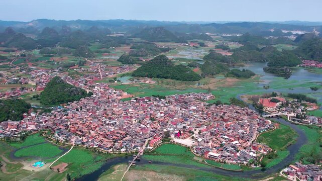 An aerial panorama of a waterside settlement surrounded by karst mountains and water bodies, with dense buildings and green fields.