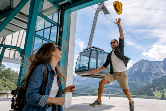 Two friends at a mountain station, a man leaps with arms raised while a woman smiles nearby as a cable car rises in the background, capturing a bright moment of adventure and joy.