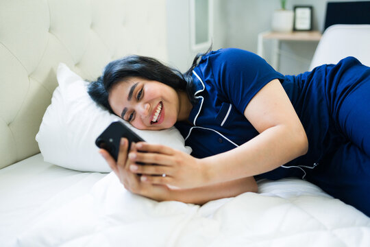 Young woman relaxing in bed wearing pajamas, smiling while using her smartphone for entertainment and communication