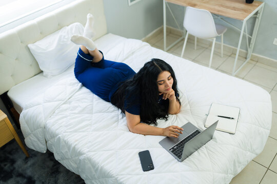 Woman working from home in pajamas, lying on bed with a laptop, enjoying a relaxed morning routine