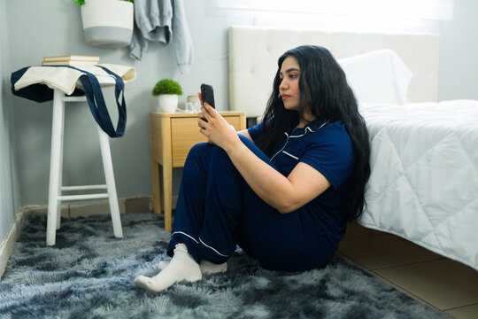 Hispanic woman feeling sad and lonely in her bedroom, sitting on the floor using a smartphone, experiencing emotional distress