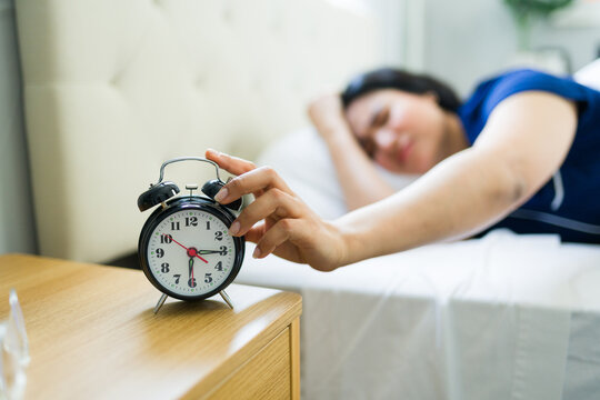 Sleepy woman waking up to a ringing alarm clock, extending hand to snooze or turn off morning alarm