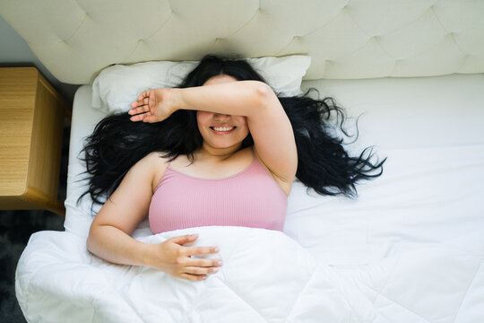High angle view of a woman waking up in bed covering face with arm, smiling after a restful night, enjoying morning light