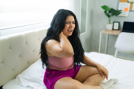Woman feeling discomfort and ache in her neck, sitting on bed with a grimace, suffering from a health issue