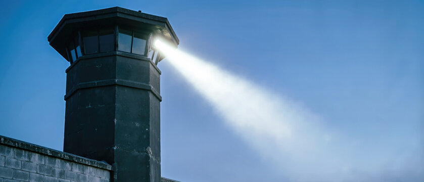 Dark concrete prison watchtower with a bright searchlight beam. Security tower and concrete wall under a deep blue sky