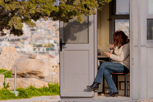 A woman sitting inside a security guard box or booth outdoors near ancient ruins, using a smartphone or device during a sunny day.