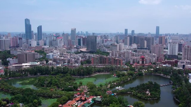 A daytime aerial panoramic view of a modern urban city, with a central park and lake surrounded by buildings of varying heights.