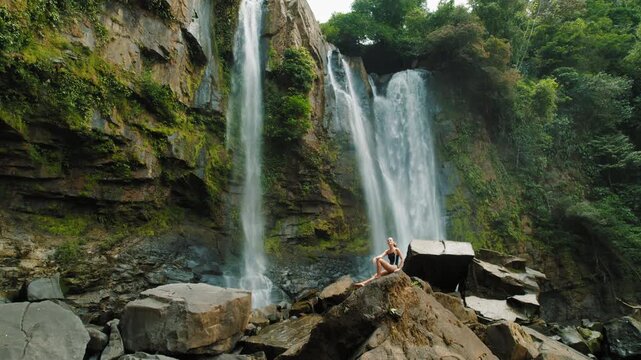 Traveler woman in swimsuit sitting on rocks near waterfalls in Costa Rica