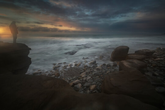 man standing on a cliff watching the sunset over rocky beach with long exposure waves in azkorri getxo basque country