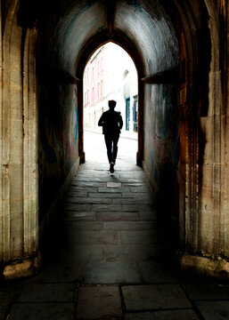 A silhouette of a person running through a dark, arched stone tunnel towards a bright city street, capturing a sense of transition and forward momentum.