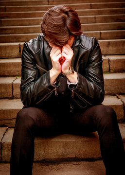 A young man in a black leather jacket sits alone on outdoor stone steps, head bowed and hands covering his face, capturing a poignant moment of deep reflection or emotional exhaustion.