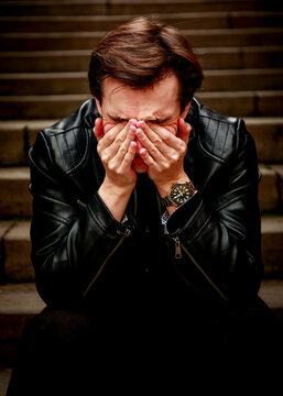 A young man wearing a black leather jacket sits alone on stone steps, covering his face with his hands in an expression of deep emotional distress, grief, or overwhelming sadness.