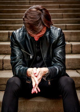 A young man wearing a black leather jacket sits alone on stone stairs with his head bowed and hands clasped, captured in a quiet moment of introspection.