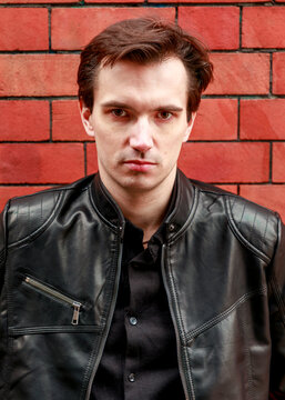 A portrait of a young man with brown hair, dressed in a black shirt and leather jacket, posing confidently against a bold red brick wall background.