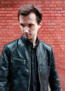 A young man in a black leather jacket poses for a portrait, looking away from the camera against a textured red brick wall.