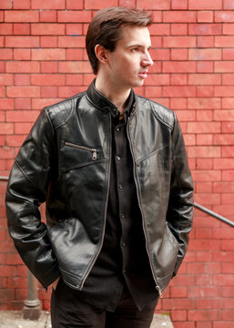 A young man poses confidently for a portrait, wearing a stylish black leather jacket over a black shirt against a textured red brick wall, showcasing a modern, casual look.