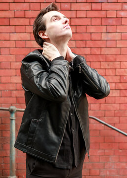 A stylish young man posing for a fashion portrait, wearing a black leather jacket against a red brick wall, gazing upwards with a contemplative expression.