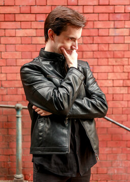 A young man in a black leather jacket stands before a red brick wall, striking a contemplative pose with his hand on his chin as he reflects on a thought.