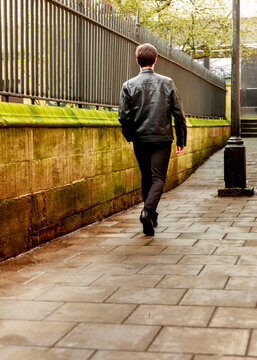 A man wearing a leather jacket and dark trousers walks along a historic stone wall with an iron fence, evoking a sense of urban movement and quiet solitude during a stroll.