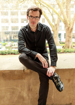 A young man wearing glasses and a black outfit sits on a stone ledge in an urban park, captured during a portrait session.