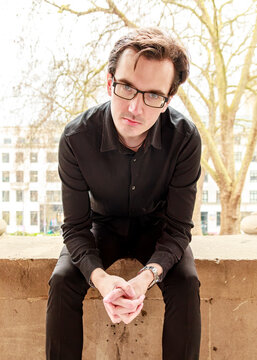 A man in a black shirt and glasses sits calmly on a stone ledge outdoors, looking directly into the camera during a portrait photography session.