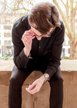 A young man in a black outfit sits on a stone wall, pausing to adjust his glasses while lost in a moment of quiet contemplation in an urban setting.