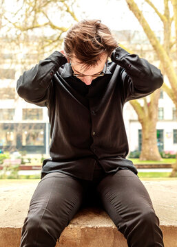 A young man in a black shirt sits outdoors with his hands behind his head and his gaze cast downward, seemingly lost in quiet contemplation or taking a moment to rest in the park.