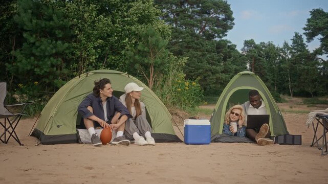 Multiethnic young adults camping on beach, sitting by tents with African American man working on laptop. Video is suitable for digital nomad, remote work, and outdoor leisure marketing