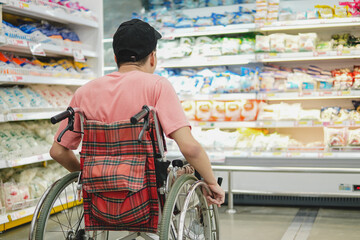 Obraz premium Back view of a young Asian man in a wheelchair looking at products on supermarket shelves, concept of accessibility, independent lifestyle, and inclusion.