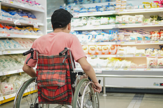 Back view of a young Asian man in a wheelchair looking at products on supermarket shelves, concept of accessibility, independent lifestyle, and inclusion.