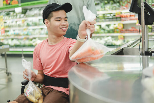 Happy young Asian man in a wheelchair placing a bag of fresh carrots on the weighing counter at the supermarket, concept of inclusive lifestyle and accessibility.