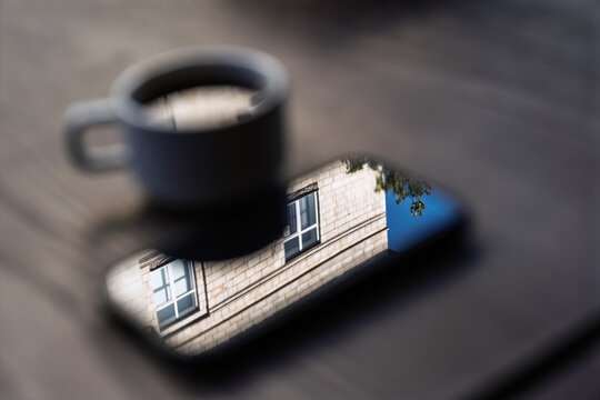 Reflection of a building facade in a smartphone screen next to a cup of coffee on a wooden table