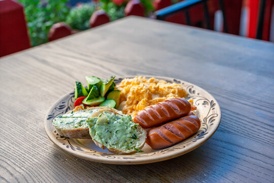 Breakfast plate with scrambled eggs, grilled sausages, bread with herb butter and fresh cucumber salad on a wooden table