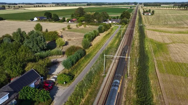 Aerial view of a passenger train traveling through the Dutch countryside, surrounded by farmland, rural homes, green fields, railway tracks, and a scenic agricultural landscape.