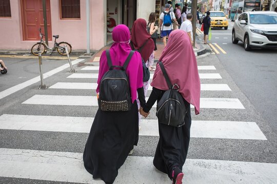 Two women wearing hijab and backpacks hold hands while crossing a city crosswalk, with pedestrians and traffic passing in the background