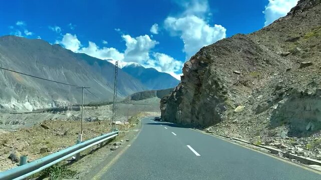 Fast-forward travel video along the Karakoram Highway (KKH) in northern Pakistan, featuring a smooth mountain road surrounded by dramatic barren valleys, rugged peaks, distant snow-covered mountains, 