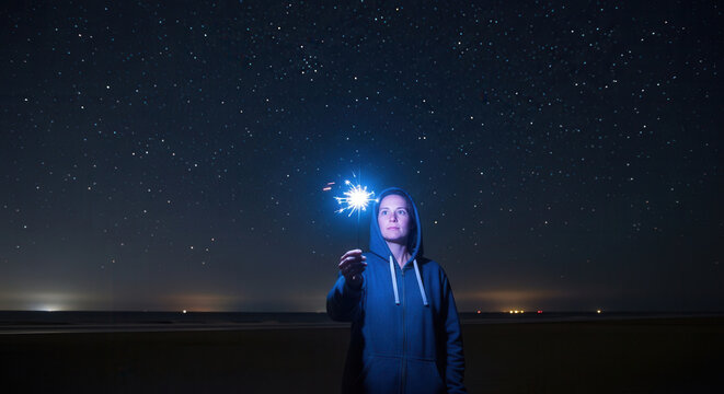 A person standing under a starry night sky holding a glowing sparkler, creating a magical and dreamy atmosphere with celestial lights in the background, evoking feelings of wonder and tranquility.