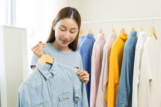 Choose clothes to wear, attractive asian young woman, girl try on appare, happy choice of shirt, female outfit on hanger in wardrobe in room closet at home. Deciding blouse what to put on which one