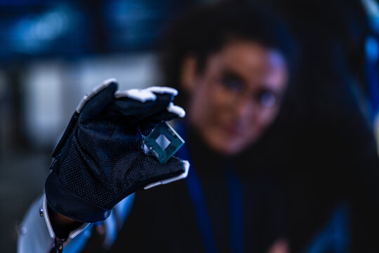 Gloved hand presenting a microprocessor chip close to camera in a dark blue-lit cybersecurity lab with security dashboards glowing in the blurred background.