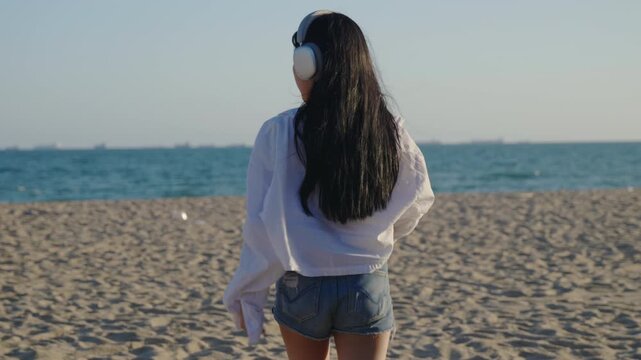 Happy woman on summer beach loves to listen to music and dance wearing wireless headphones, white shirt and denim shorts, back view of female enjoying sunny day near ocean.