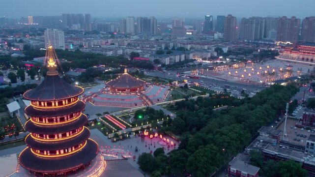 Aerial view of illuminated traditional pagoda and adjacent square, set in modern urban area at dusk.