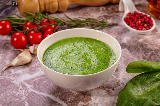 Creamy spinach soup in white bowl with fresh ingredients on marble surface