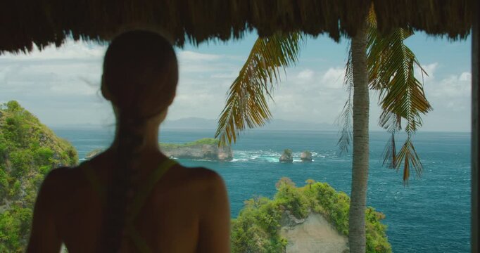 Back view of woman watching ocean waves from rustic wooden hut balcony in Nusa Penida Bali island