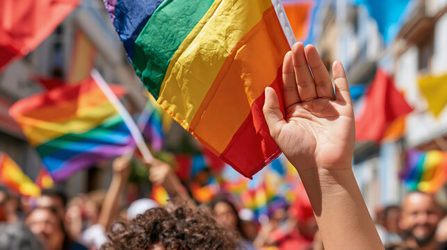 Hand holding small rainbow flag at pride parade with crowd and colorful flags in background celebrating lgbtq rights